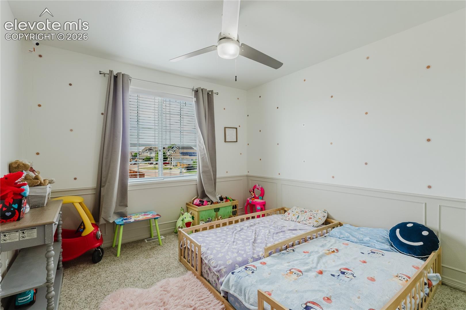 Carpeted room featuring a ceiling fan with light fixture, window with blinds and drapes, and wainscoting