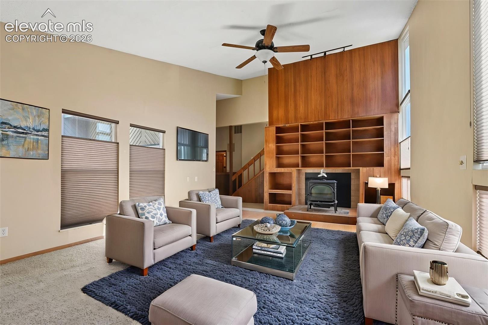 Vaulted ceilings, floor-to-ceiling wood-panelled accent wall, built-in wooden shelving, freestanding wood-burning stove, and expansive vertical windows