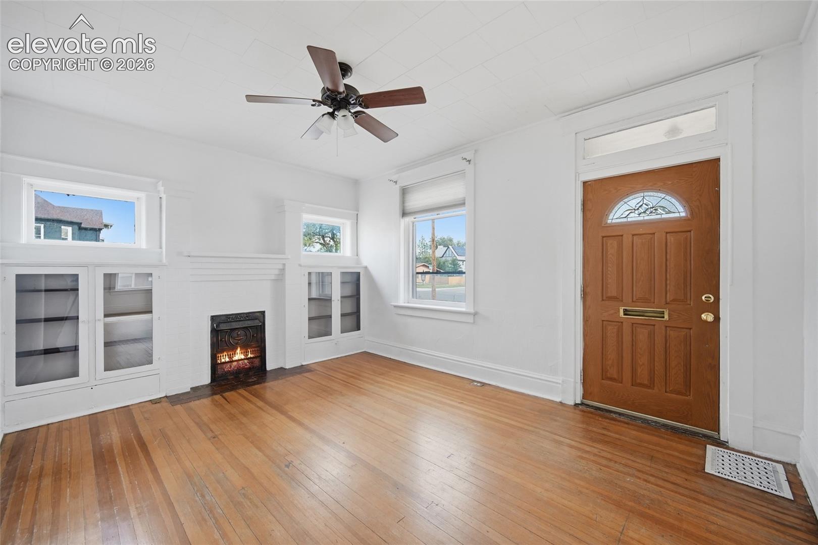 Wood-finish flooring, built-in shelving flanking a fireplace, ceiling fan with integrated lighting, and wooden entry door featuring a transom window
