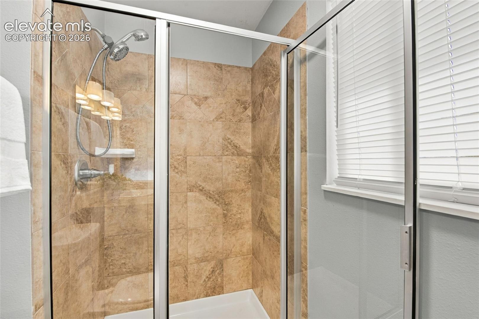 Walk-in shower featuring glass paneling, floor-to-ceiling stone-tile surround, adjustable handheld showerhead, and window with built-in blinds