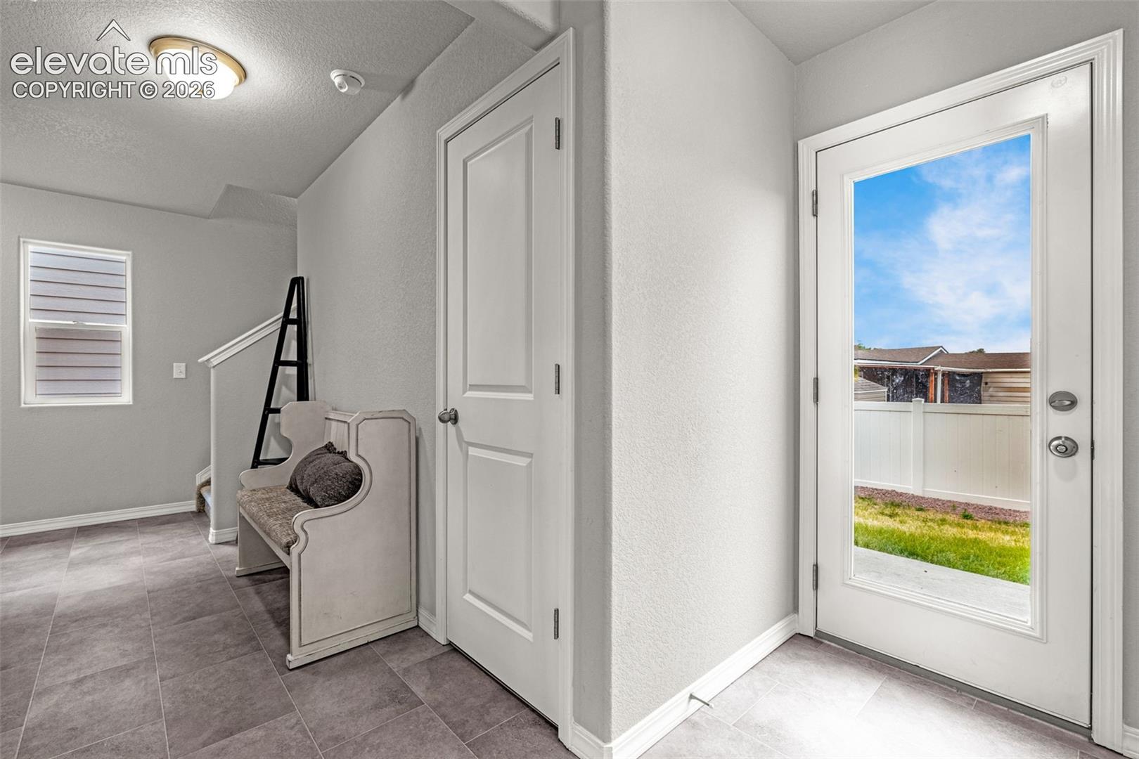 Entryway featuring a full-light exterior door, grey tile flooring, neutral wall tones, white baseboards, and a window with horizontal blinds
