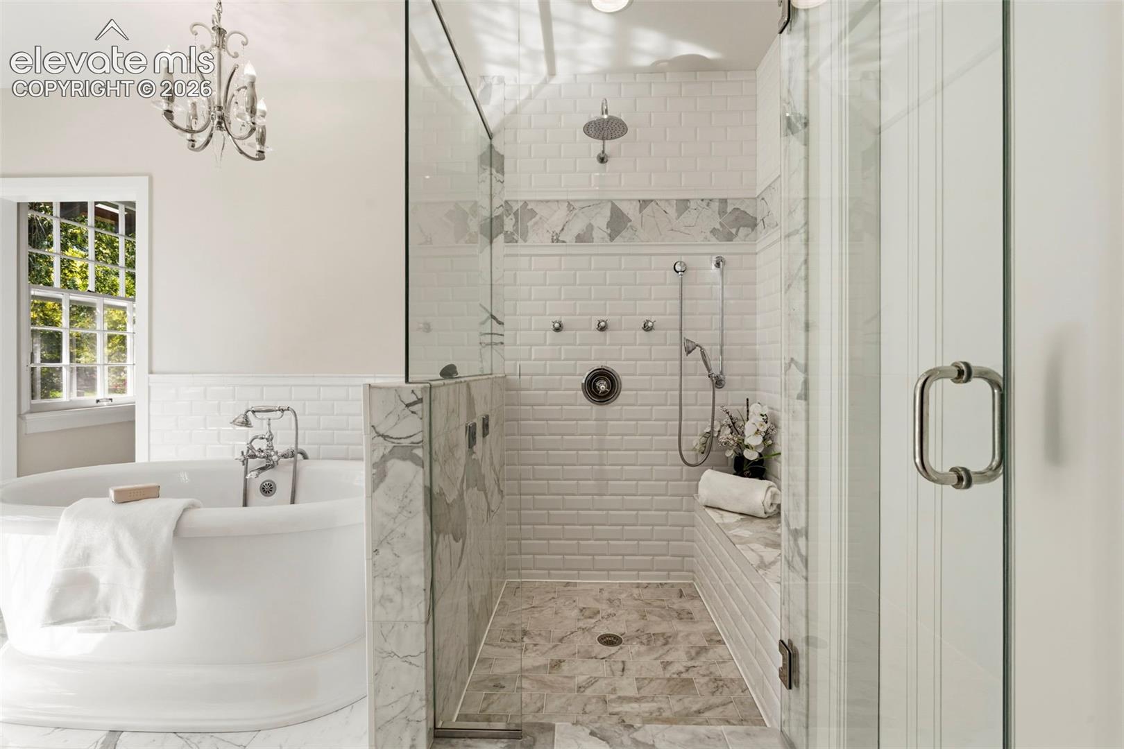 Freestanding soaking tub with polished chrome faucet, glass-enclosed walk-in shower featuring subway tile walls, stone tile flooring, and integrated shower bench