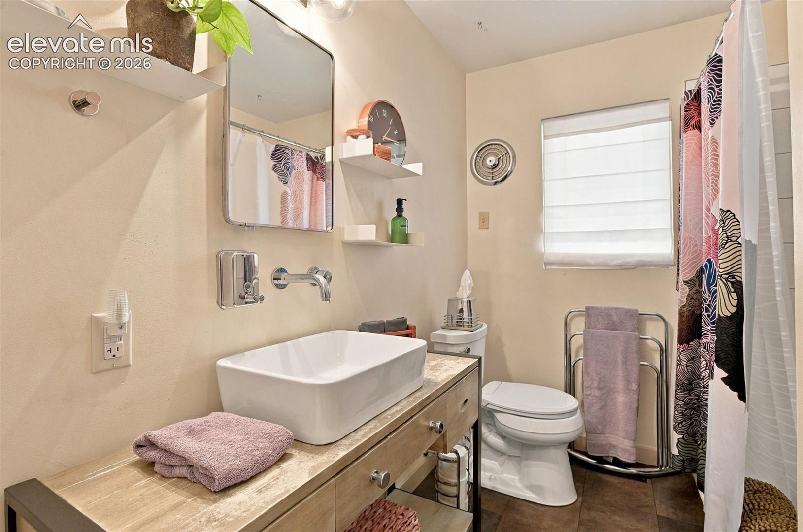 Bathroom featuring a wood-finish vanity, rectangular vessel sink, wall-mounted faucet, rectangular wall mirror, and built-in wall shelving