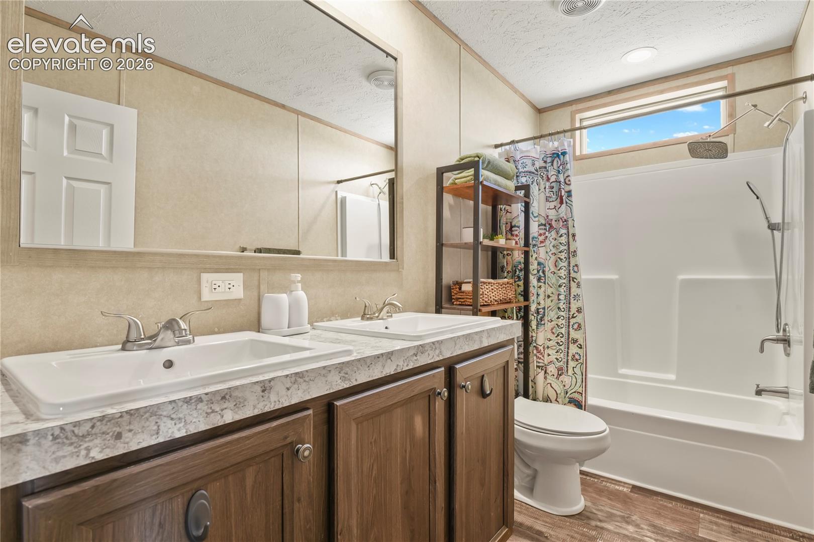Dual-sink vanity with a stone-finish countertop, full-width wall mirror, wood-finish cabinetry, and a combination bathtub and shower enclosure with a transom window