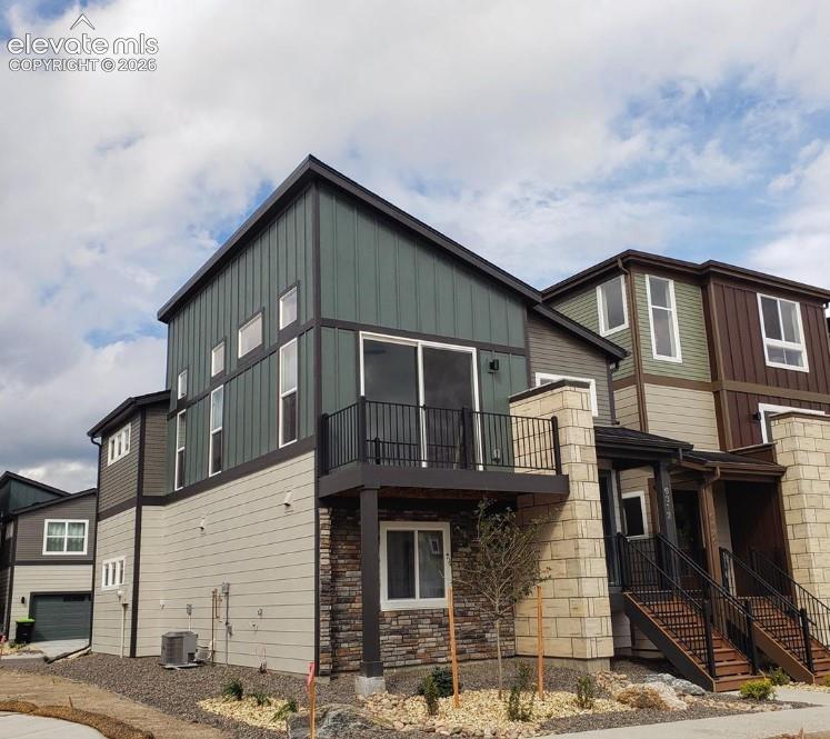 Multi-story exterior featuring vertical metal siding, horizontal lap siding, stacked stone veneer accents, an elevated balcony with a black metal railing, and an exterior staircase