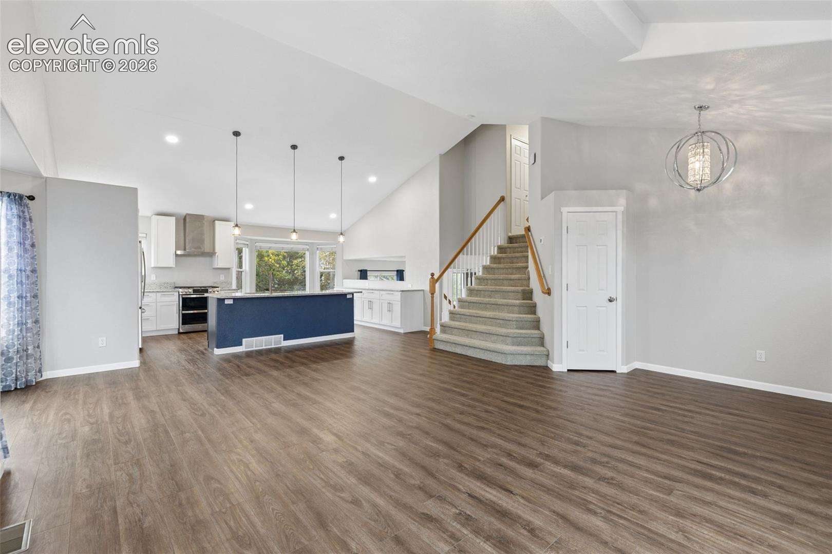 Open-concept interior featuring vaulted ceilings, wood-finish flooring, a kitchen island with pendant lighting, and a staircase with a wooden handrail