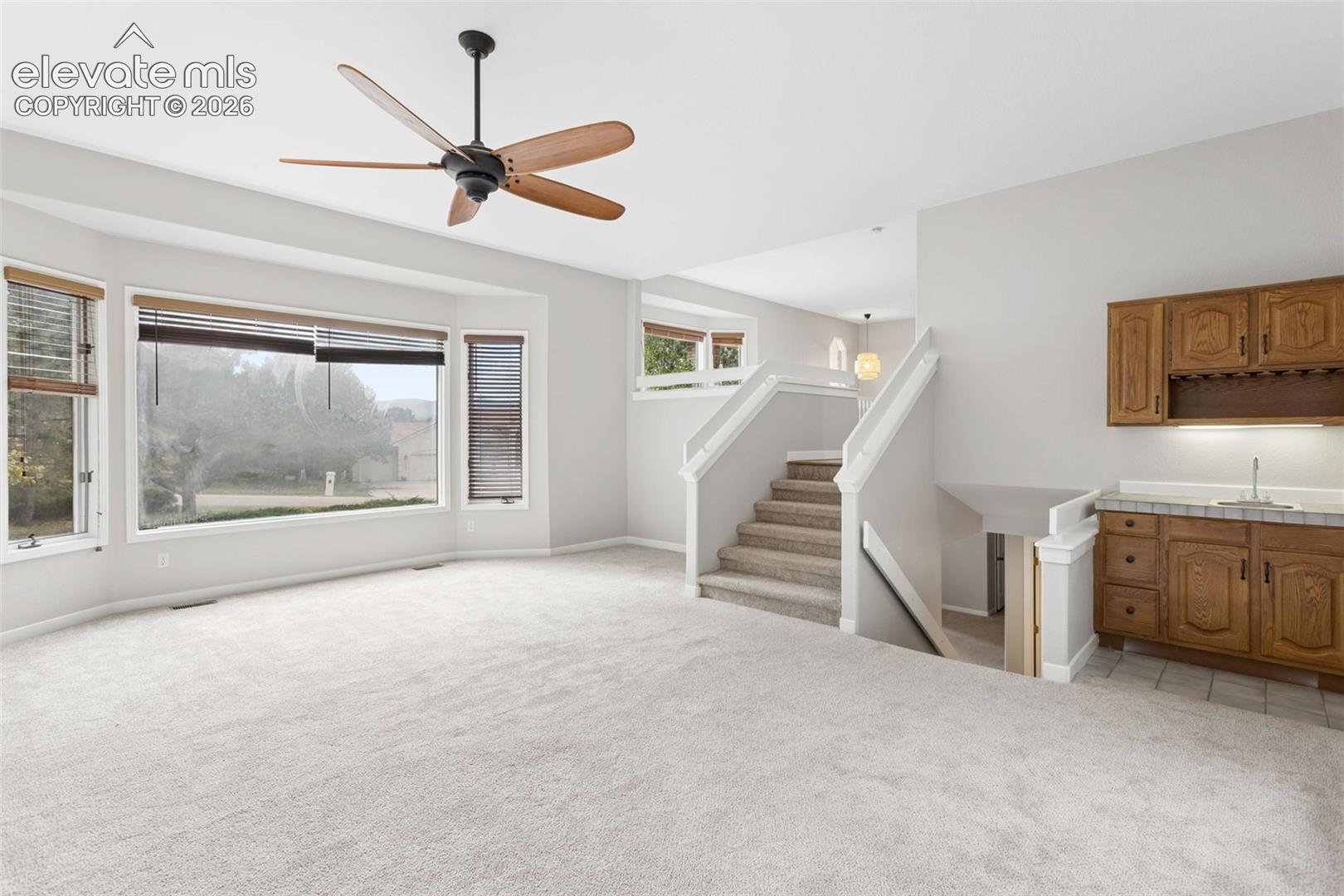 Expansive carpeted living space with a ceiling fan, a bay window, and a built-in wet bar with wood cabinetry