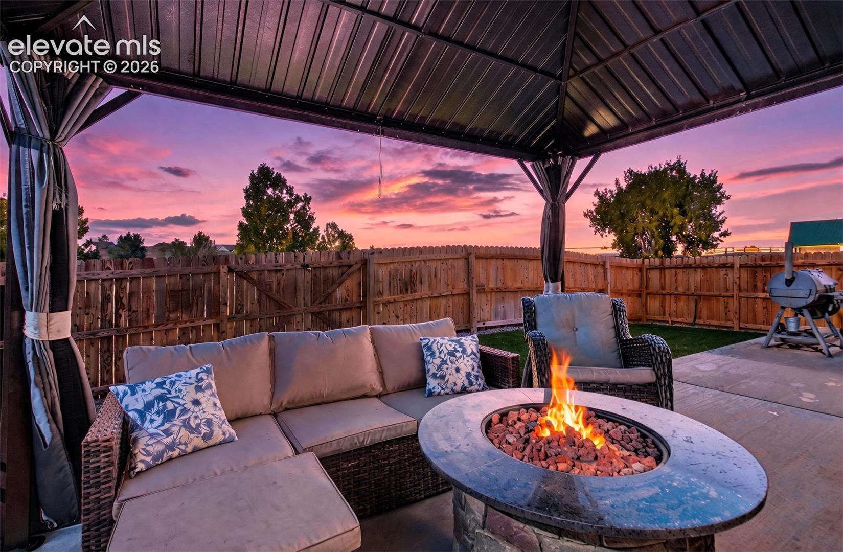 Covered patio featuring a metal gazebo structure, stone fire pit, and concrete hardscaping enclosed by a wooden privacy fence