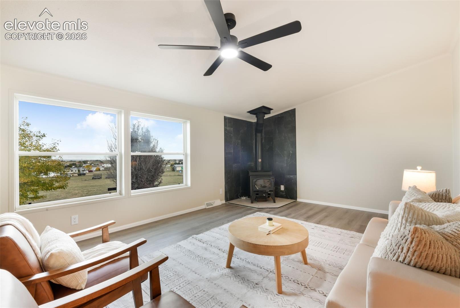 Wood-finish flooring, accent wall with a built-in stove, two expansive windows, and a ceiling fan with integrated lighting