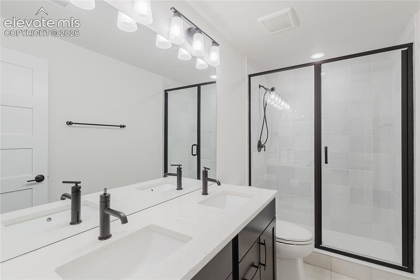 Bathroom in the lower level with dual integrated sinks and black-finish fixtures, ample mirror, glass-enclosed shower with black framing, and white tiled walls
