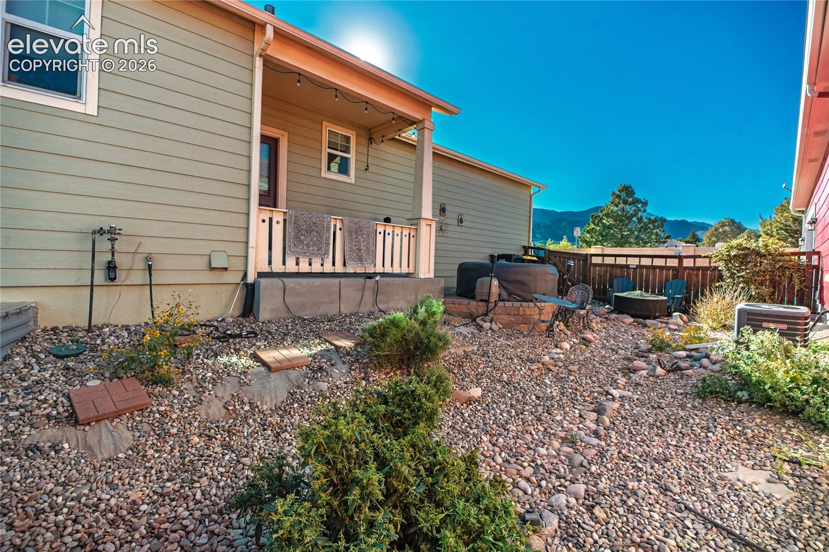 Rear exterior featuring horizontal lap siding, a covered porch with a wood balustrade, xeriscaped grounds with decorative stone, a raised stone fire pit, and mountain scenery