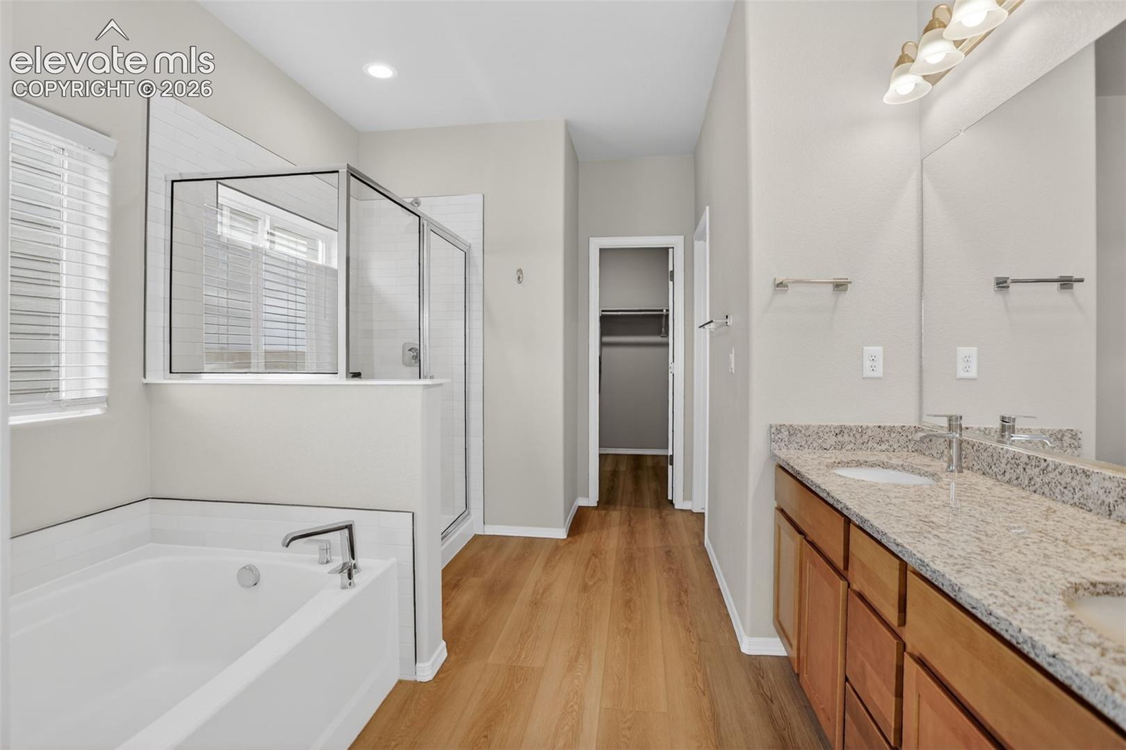 Bathroom with wood-finish flooring, dual vanity featuring granite countertops and undermount sinks, and a walk-in shower with a glass enclosure
