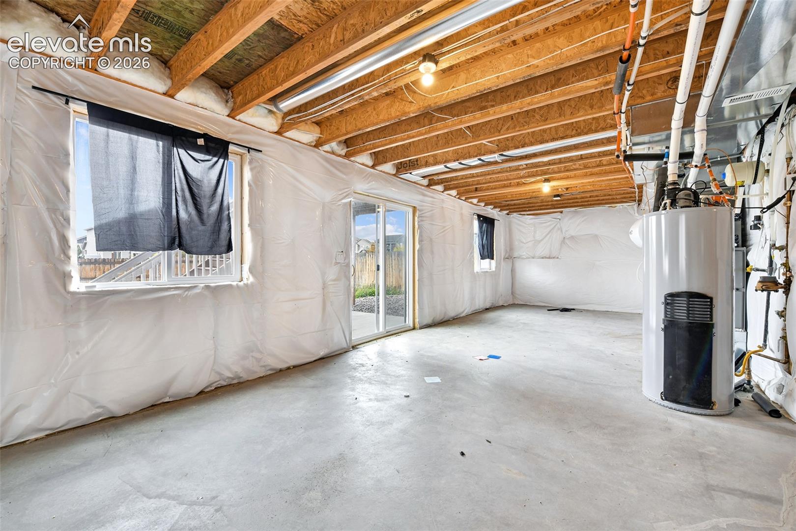 Unfinished basement area featuring exposed timber ceiling joists, concrete flooring, sliding glass door, and vinyl-sheathed foundation walls