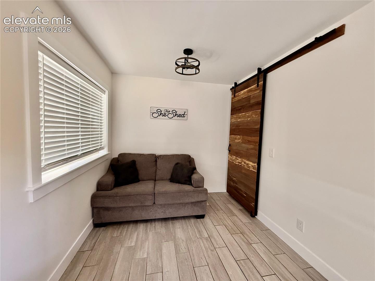 Wood-finish tile flooring, a large window with blinds, a ceiling-mounted light fixture, and a wood barn door with a black metal track