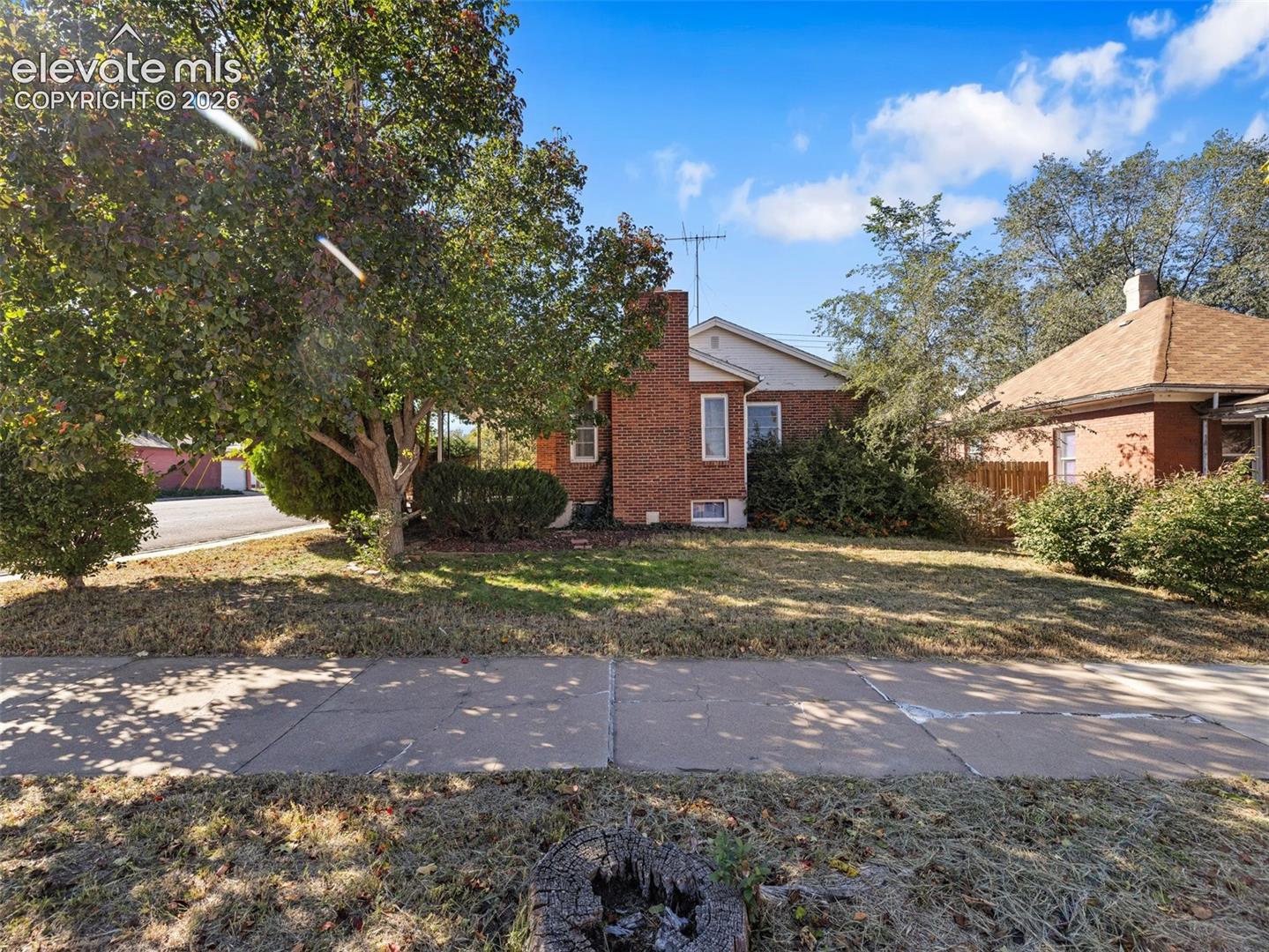 Brick exterior featuring a chimney, asphalt shingle roof, mature landscaping, and a concrete sidewalk