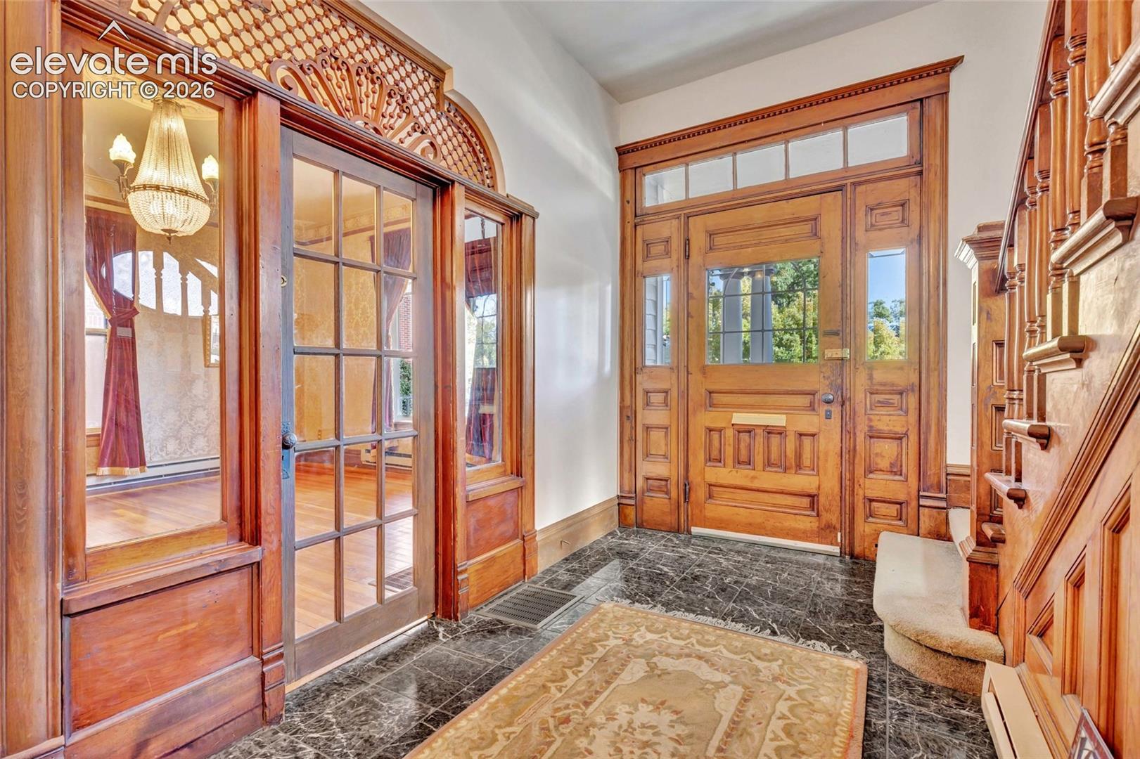Entryway featuring natural wood trim, a paneled wood door with a transom window, ornate decorative millwork, a polished stone floor, and a wooden staircase