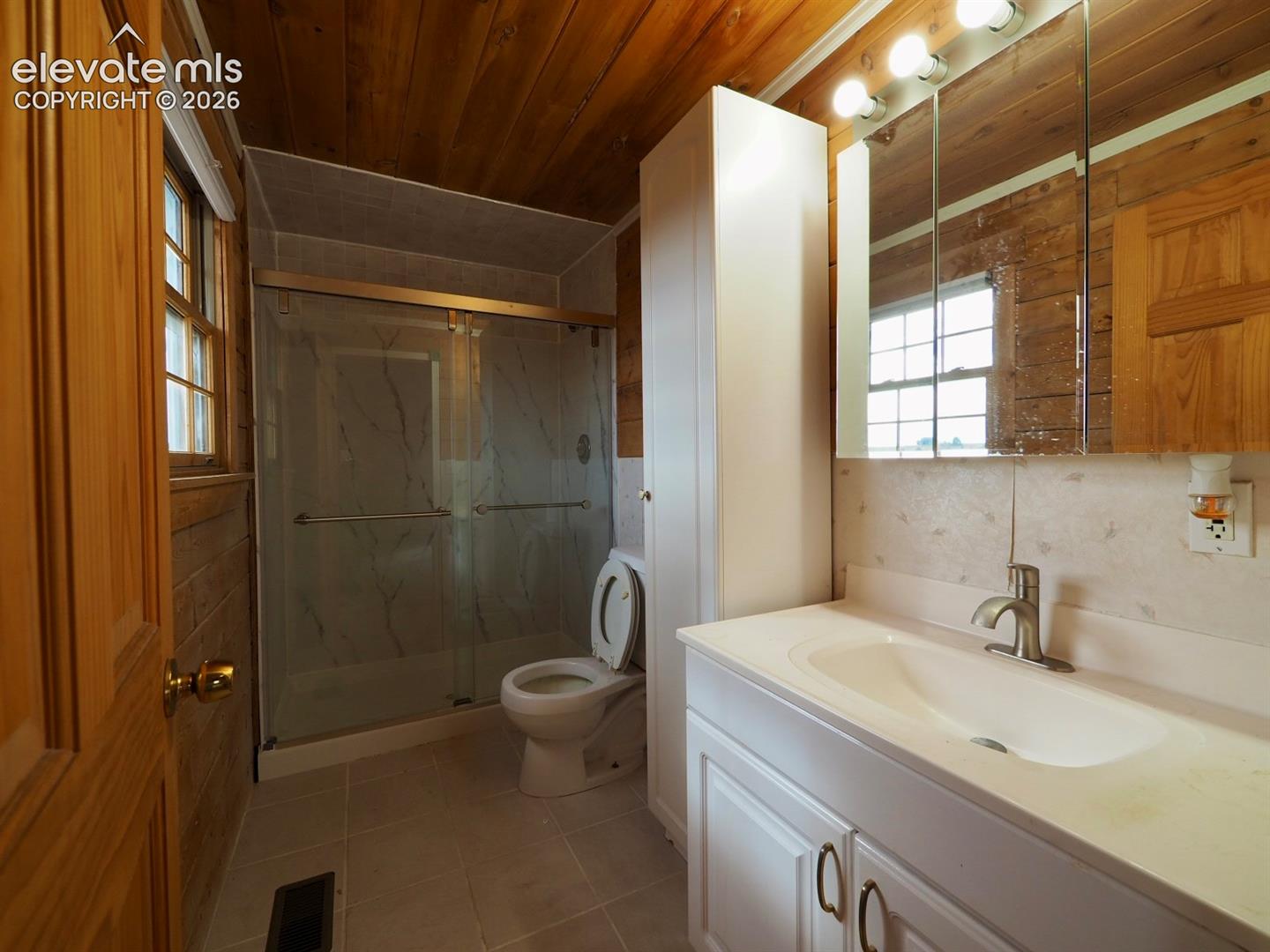 Bathroom featuring wood paneling on the walls and ceiling, a walk-in glass-enclosed shower, a built-in floor-to-ceiling storage cabinet, and a vanity with a white countertop