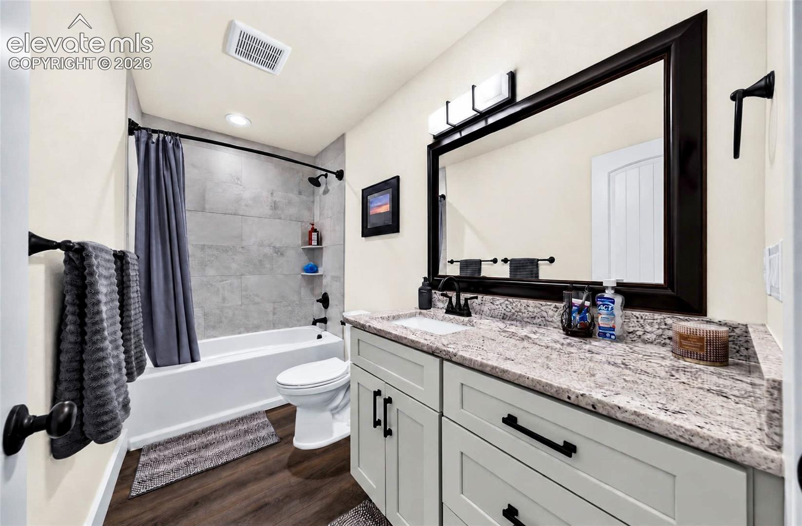 Bathroom featuring light-colored cabinetry with dark hardware, stone-finish countertop, rectangular undermount sink, framed wall mirror, and tub-shower combination with tile surround
