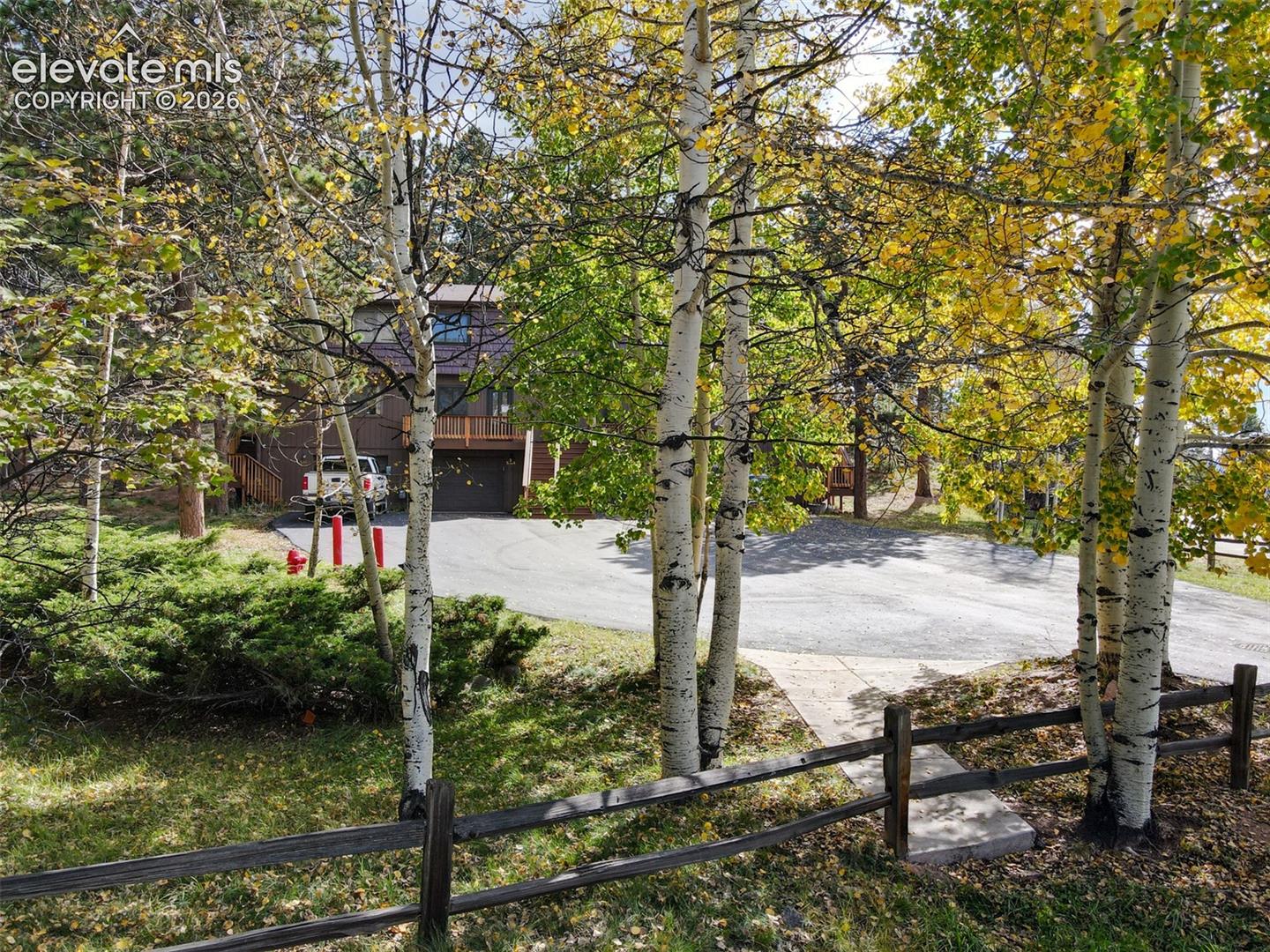 Split-rail fence framing the property, mature deciduous trees, evergreen shrubs, and an asphalt driveway, with a multi-level exterior and attached balcony