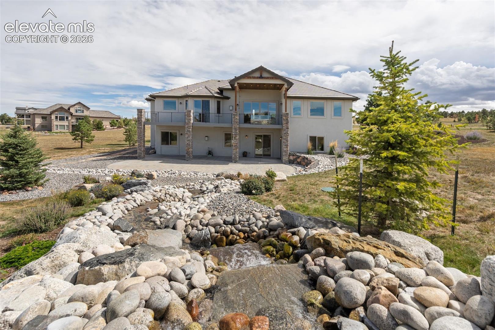 Stucco exterior with stone accents, multiple balconies, a lower-level patio, and a natural rock water feature