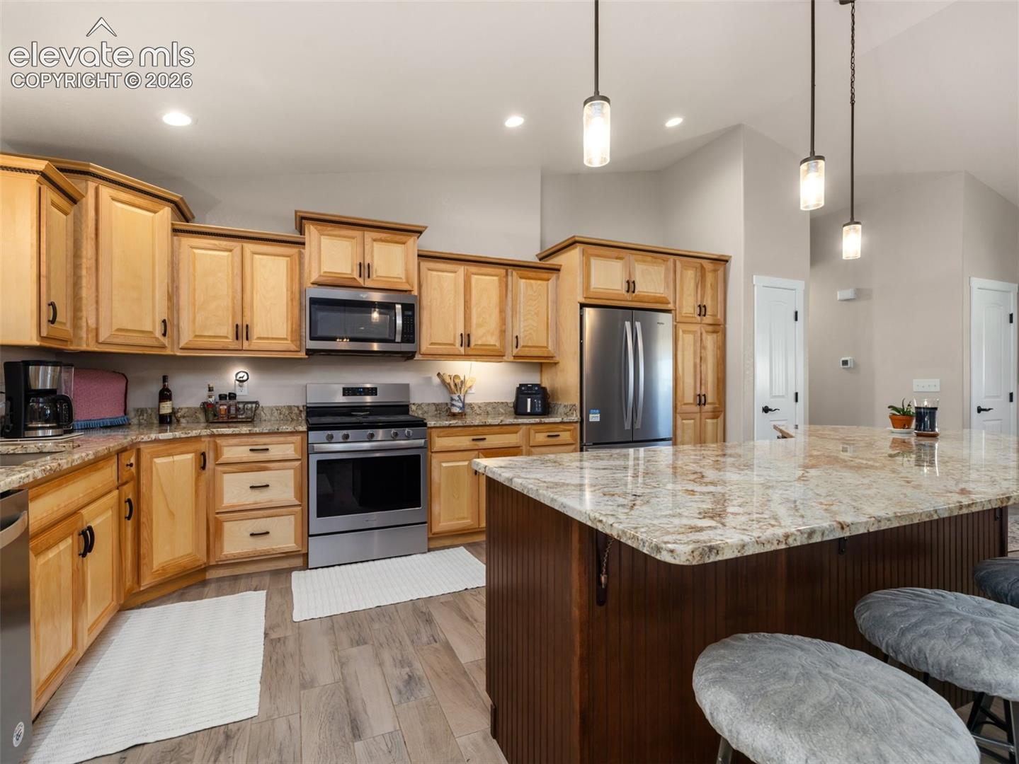 Wood-finish flooring, stainless steel appliances, recessed lighting, pendant lighting, and a kitchen island with stone countertops