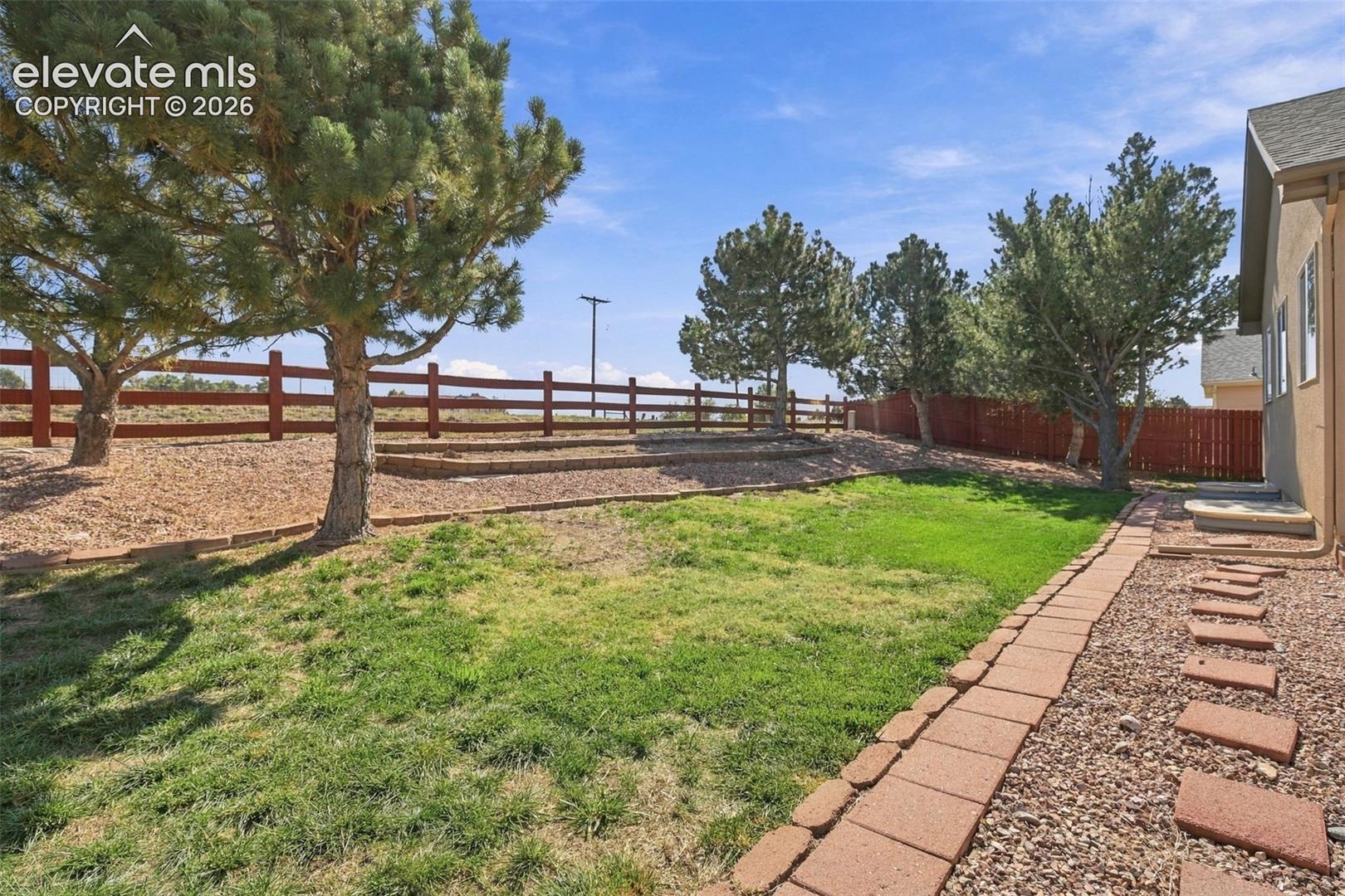 Expansive tiered backyard featuring a grass lawn, mature trees, a red wood fence, a flagstone path, and a gravel bed