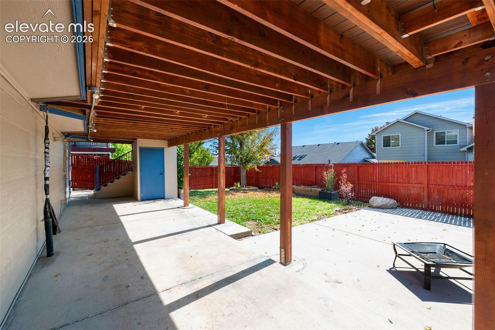 Covered patio with exposed wood beams, concrete flooring, private storage area with blue door, and red privacy fence