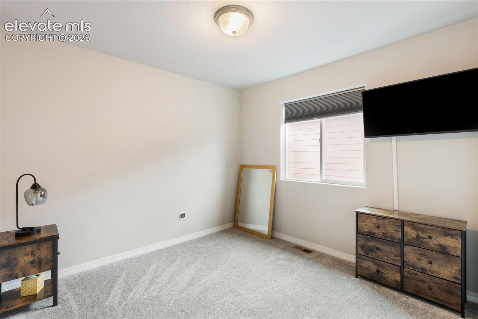 Bedroom featuring a window with a roller shade, light-colored carpeting, a flush-mount ceiling light fixture, and white baseboards