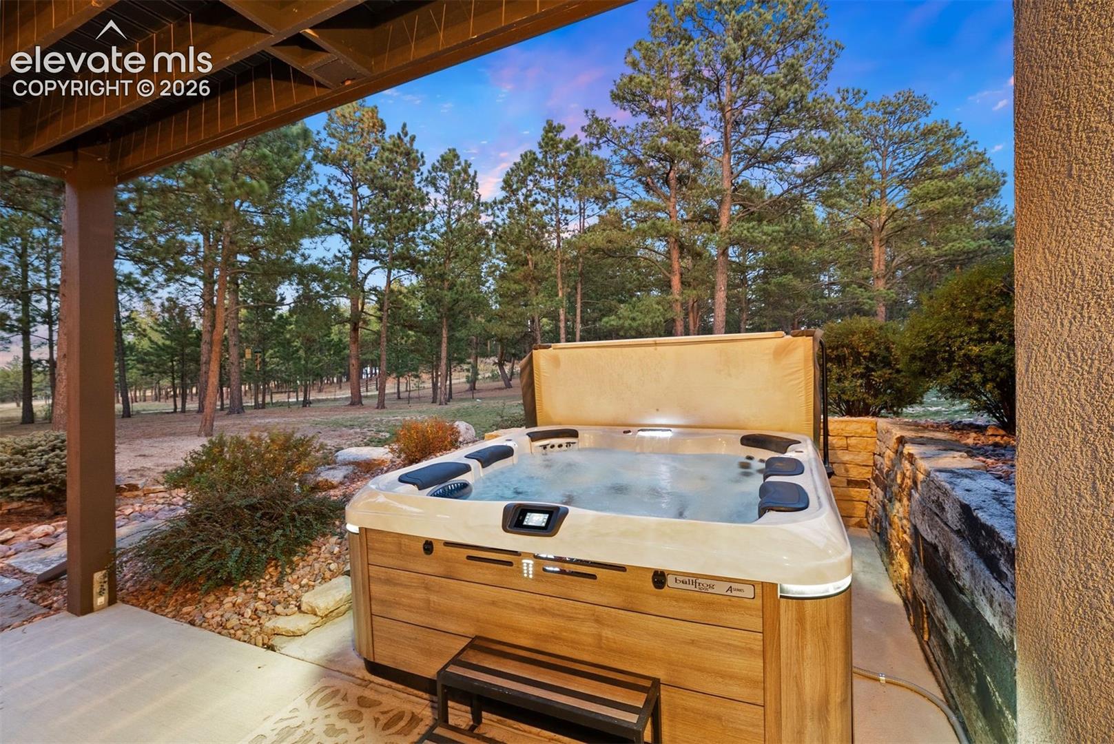 Covered patio featuring an integrated spa, wood-finish siding, natural stone retaining wall, and surrounding pine forest