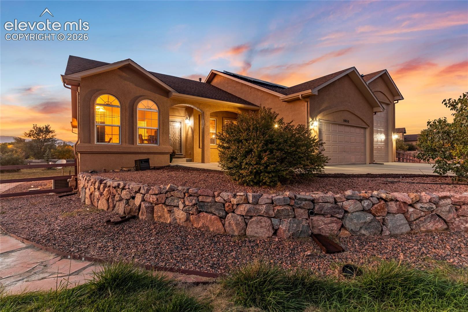 Stucco exterior featuring arched windows, a multi-car garage, and a natural stone retaining wall with low maintenance xeriscape 