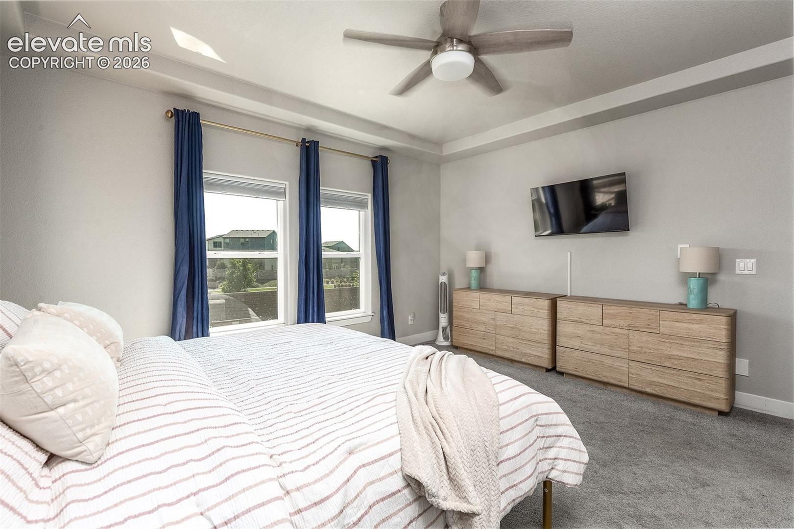 Tray ceiling with recessed lighting, two windows with blinds, ceiling fan, carpet flooring, and wall-mounted television