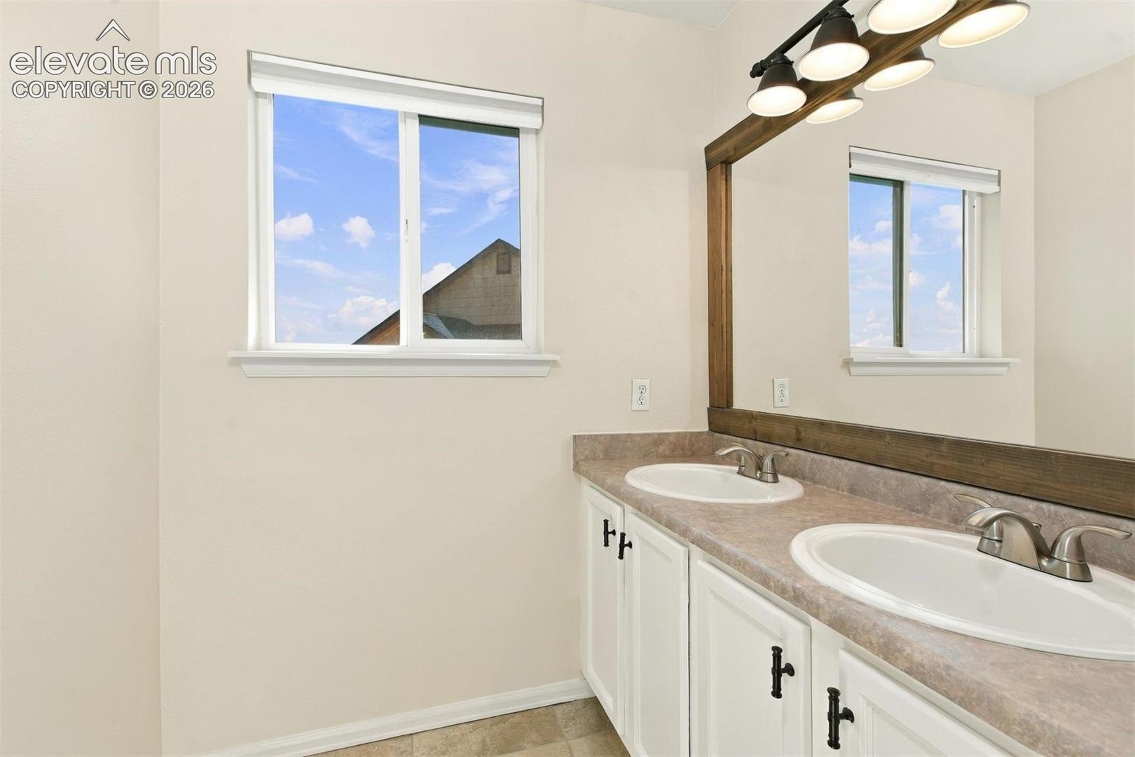 Dual-sink vanity with matte-finish countertop, white cabinetry with dark hardware, large wood-framed mirror, multi-bulb overhead light fixture, and windows with integrated blinds