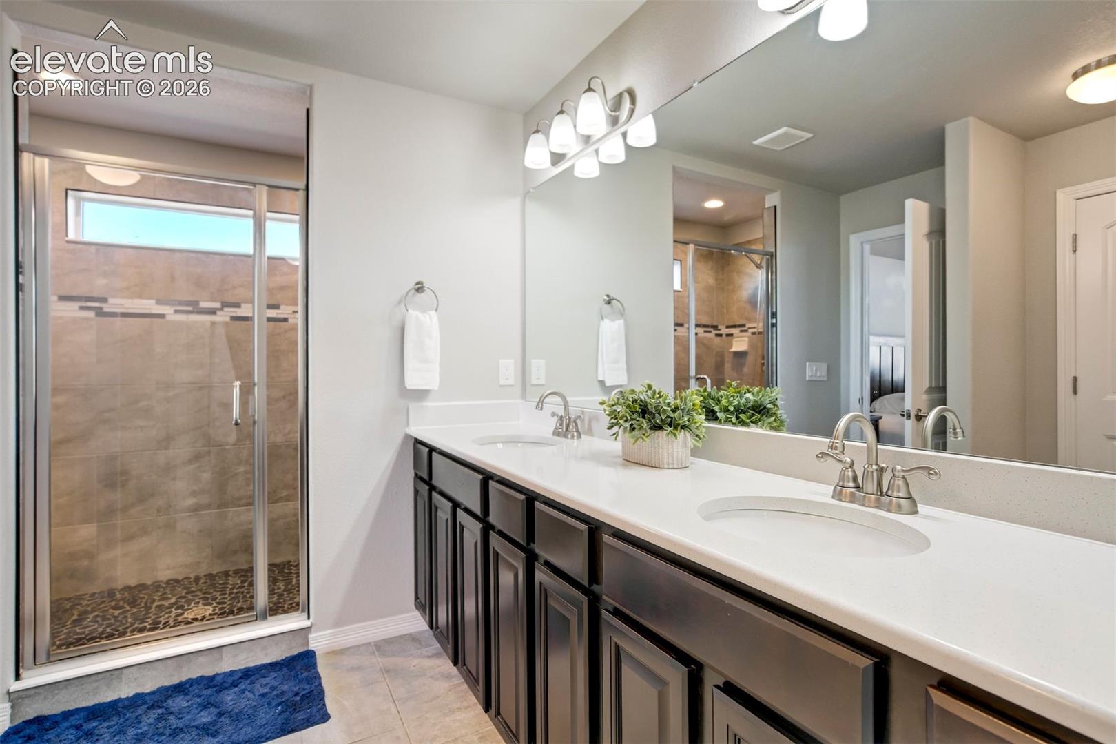 Double vanity with undermount sinks and chrome faucets, large mirror, dark wood-finish cabinetry, and a walk-in shower with glass enclosure and pebble flooring