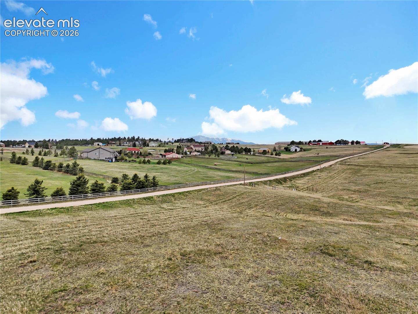 Expansive acreage featuring rolling grass terrain, mountain backdrop, and rail fencing along a gravel road