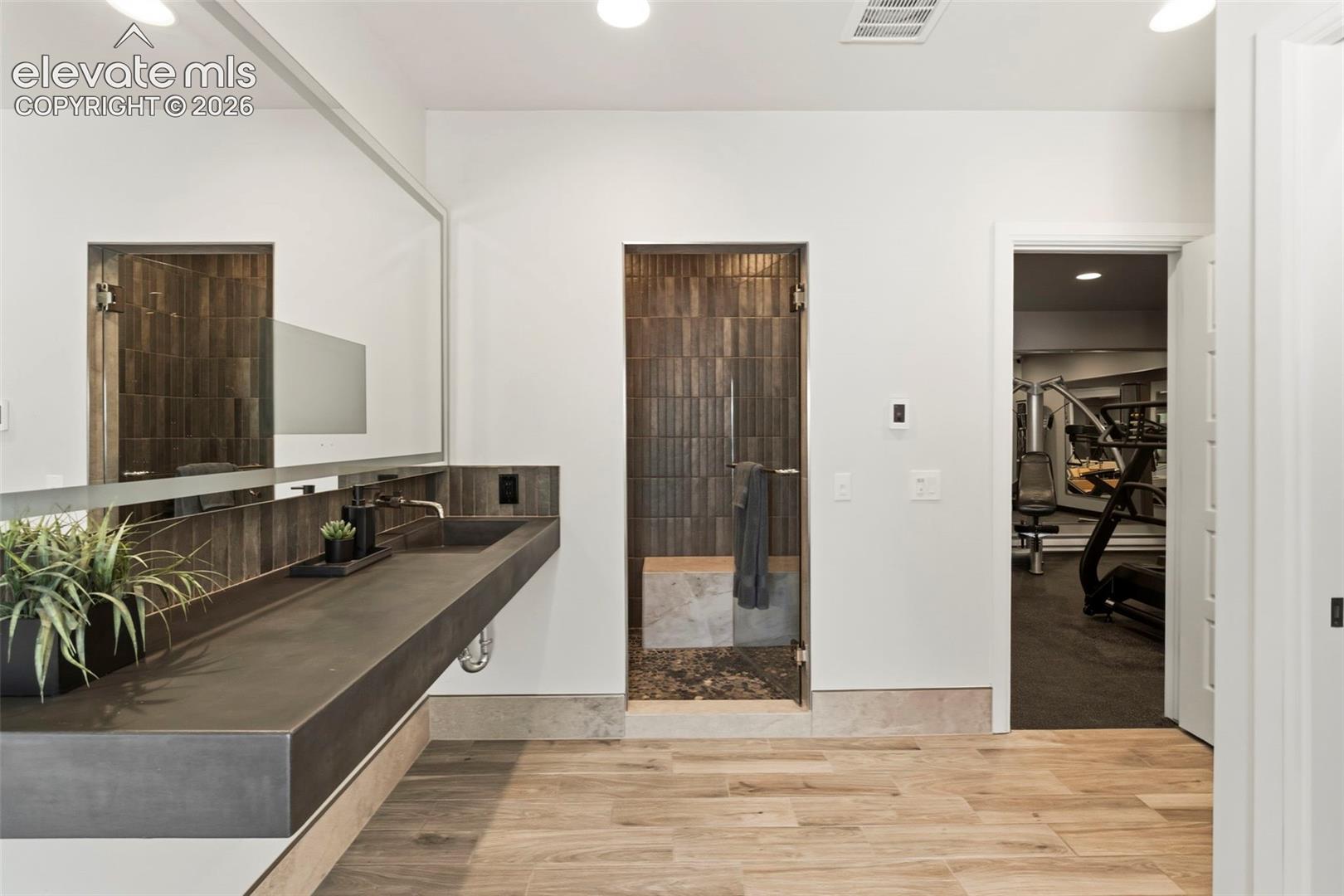 Floating vanity with a concrete-finish countertop, integrated sink, full-width mirror, tile-surround shower with a stone bench, and wood-finish flooring