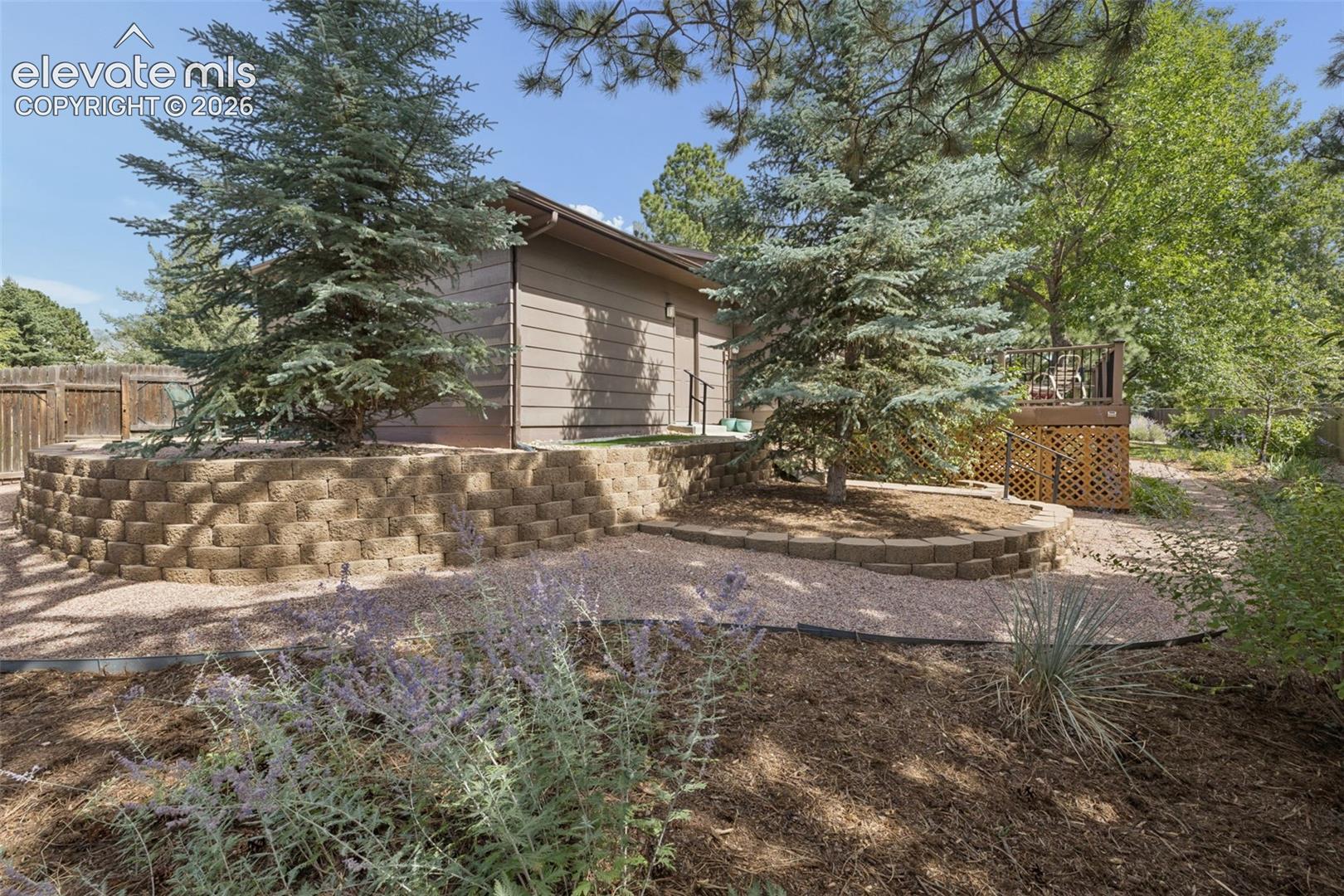 Backyard featuring a stone-retaining wall, gravel pathway, mature trees, tiered landscaping, and wood perimeter fencing