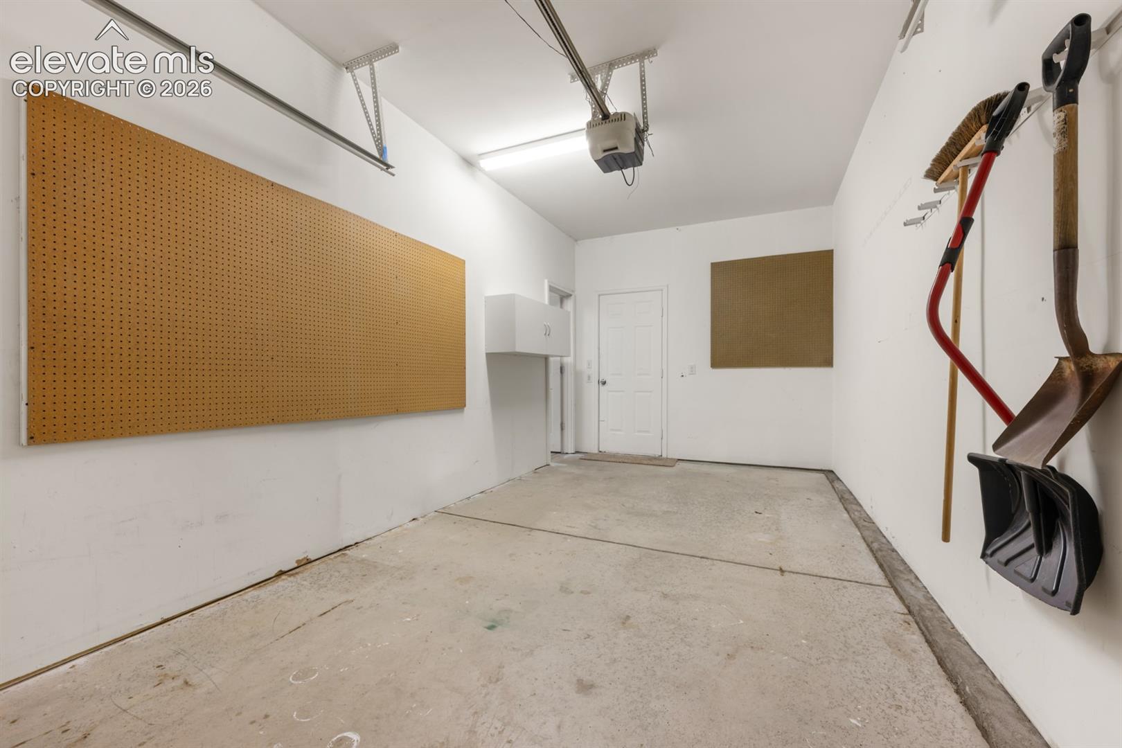 Garage featuring concrete flooring, white walls, overhead lighting, an integrated automatic garage door opener, and wall-mounted pegboard organizers