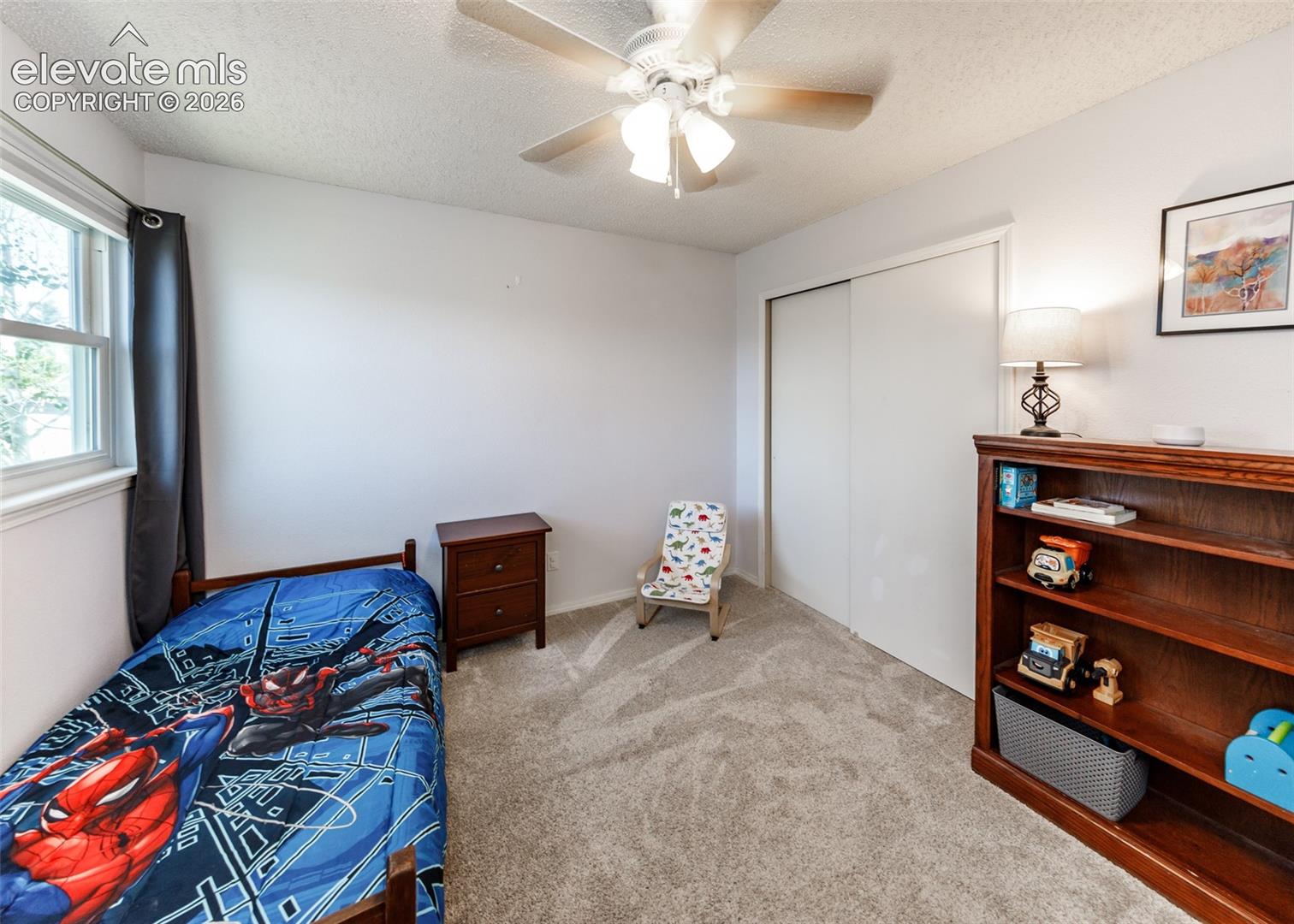 Textured carpet flooring, window with sash, ceiling fan with light fixture, built-in closet with sliding doors, built-in shelving unit