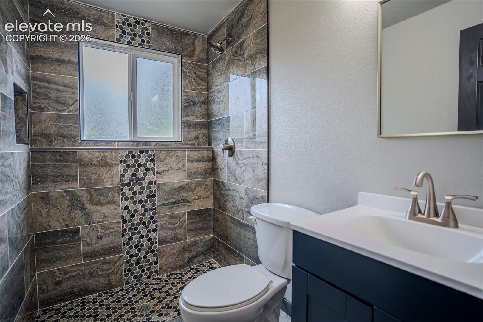 Bathroom featuring walk-in shower with large-format stone-finish tile, decorative mosaic tile inlay, built-in wall niche, dark-finish cabinetry, and solid-surface countertop with undermount sink