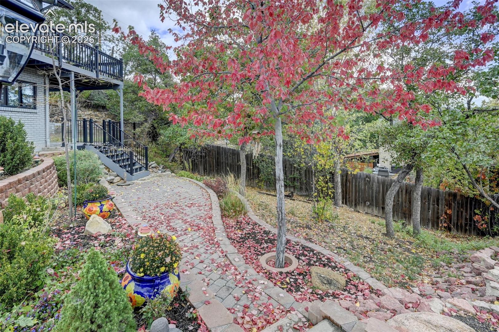 Outdoor space featuring a paver stone walkway, elevated balcony with metal railings, tiered stone retaining wall, and mature deciduous landscaping