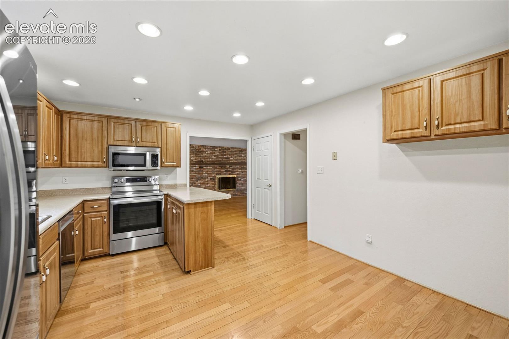 Kitchen featuring wood cabinetry, stainless steel appliances, recessed lighting, wood-finish flooring, and a brick fireplace visible in the background