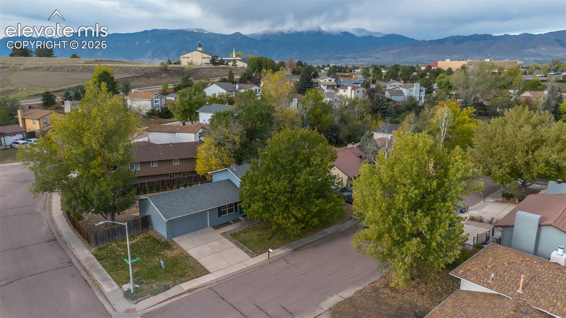 Aerial perspective of a corner lot property featuring a concrete driveway, multi-gabled shingle roof, mature deciduous trees, and a mountain range backdrop