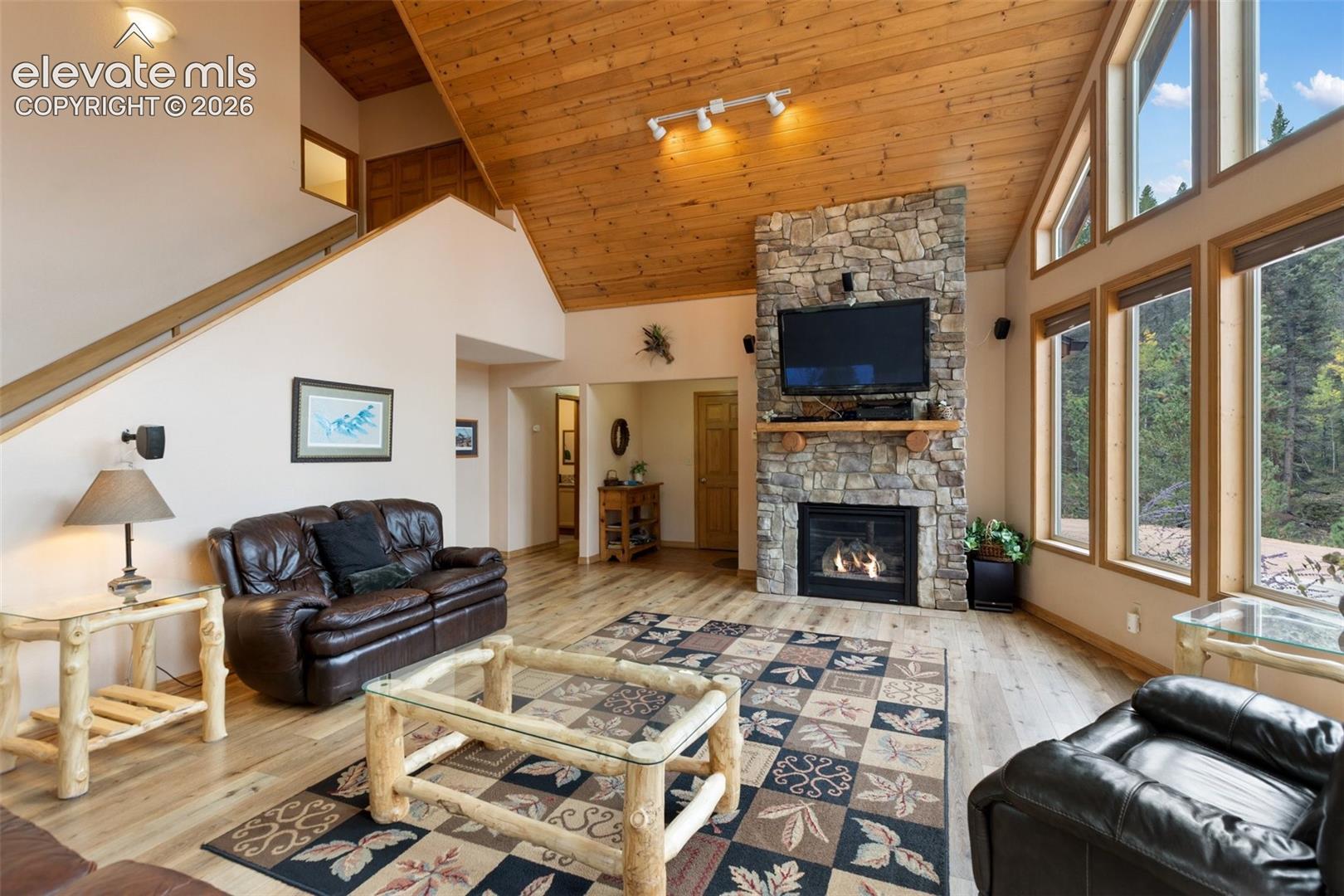 Vaulted wood-paneled ceiling, stone fireplace with mantel, hardwood flooring, large windows, and an interior staircase with wood railing