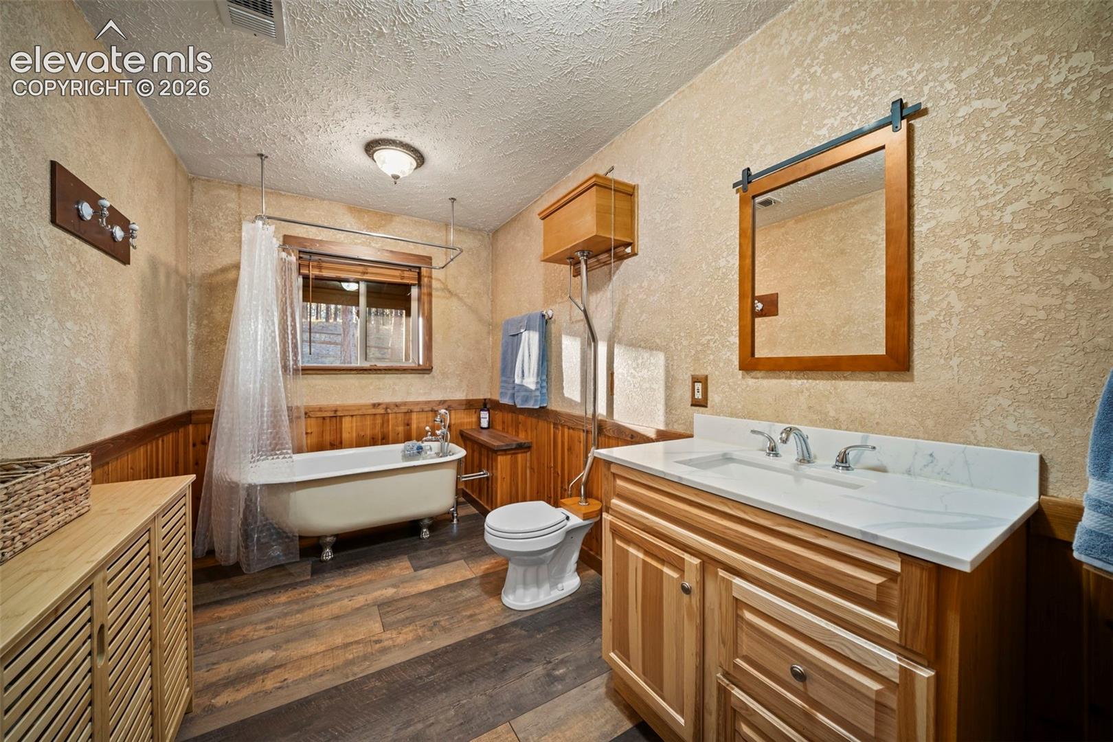 Clawfoot soaking tub, wood-finish flooring, wood wainscoting, wall-mounted mirror with sliding barn door hardware, and vanity with stone countertop