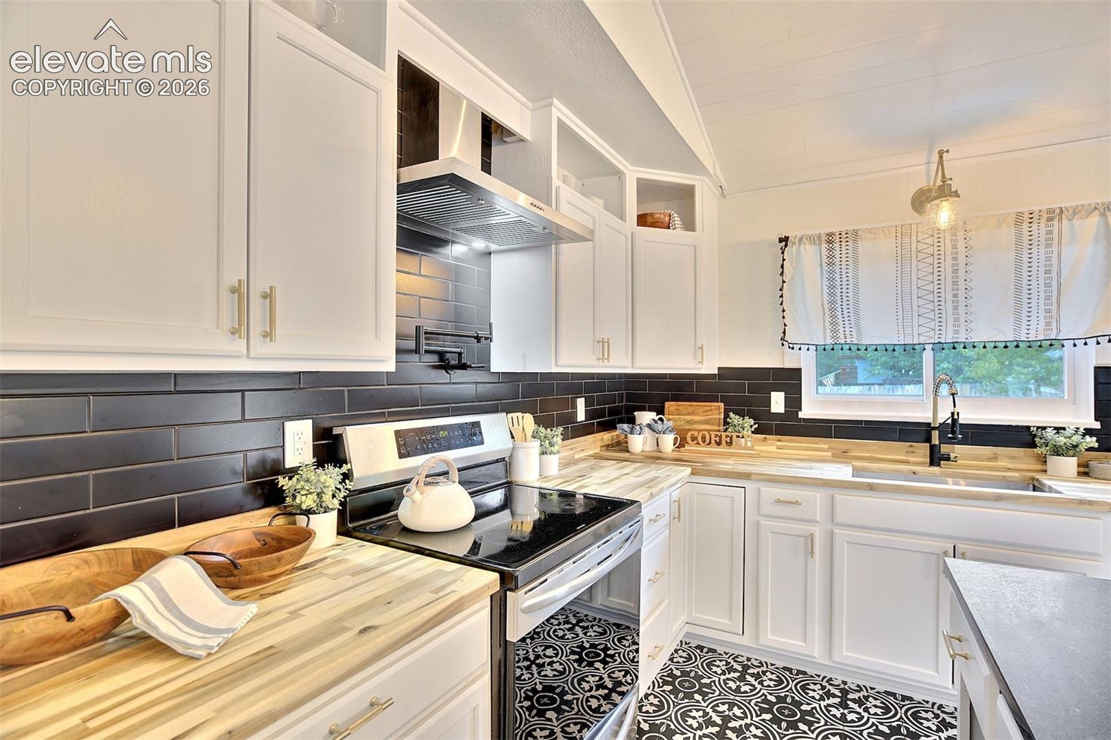Kitchen featuring white shaker-style cabinetry, black subway tile backsplash, butcher block countertops, stainless steel range with overhead hood, and patterned tile flooring