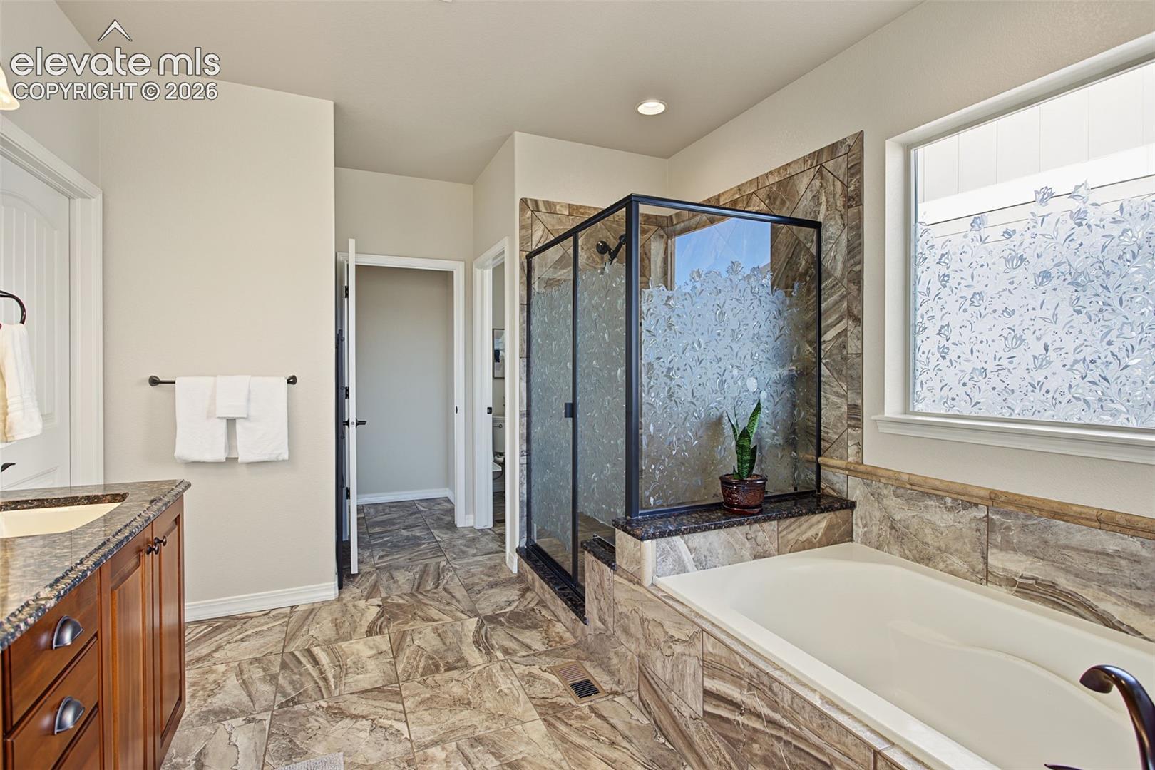 Bathroom featuring stone-finish floor tiling, a soaking tub with stone-tiled surround, a separate glass-enclosed shower with a black frame, and a wooden vanity with a stone-finish countertop