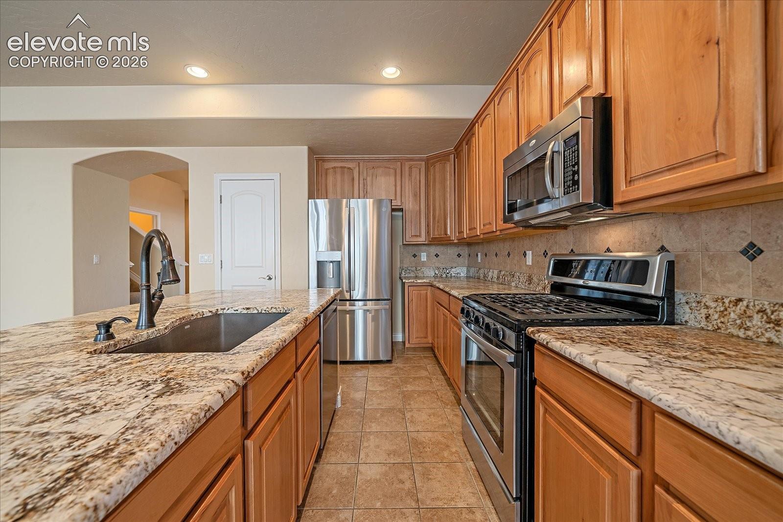 Kitchen featuring stone countertops, wooden cabinetry, stainless steel appliances, tiled backsplash, and recessed lighting