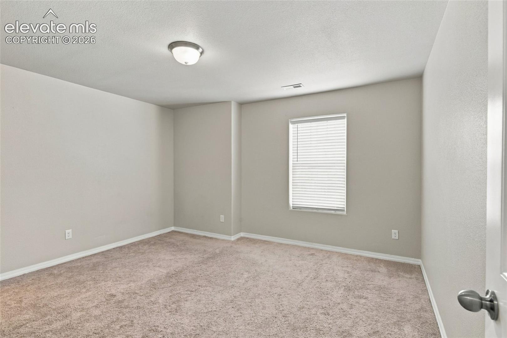 Neutral-toned interior space featuring wall-to-wall carpeting, a single-window configuration with installed blinds, and a central flush-mount ceiling light fixture