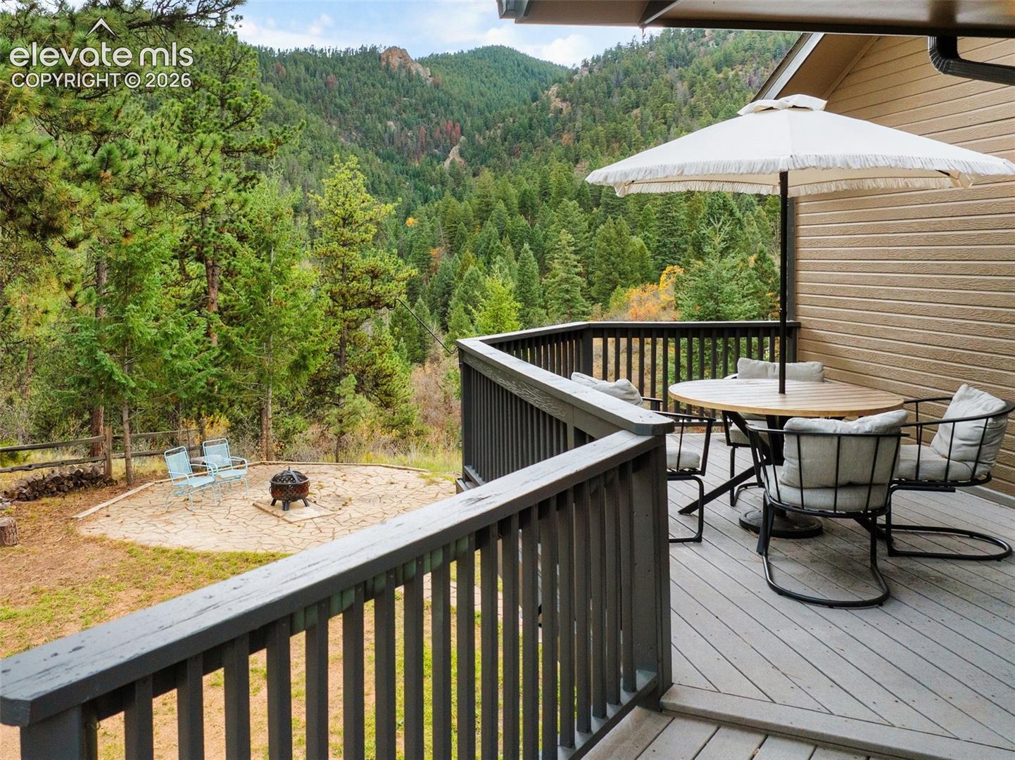 Elevated wood-finish deck with a dark-tone railing, paired with a ground-level circular stone patio set against a forested mountain backdrop