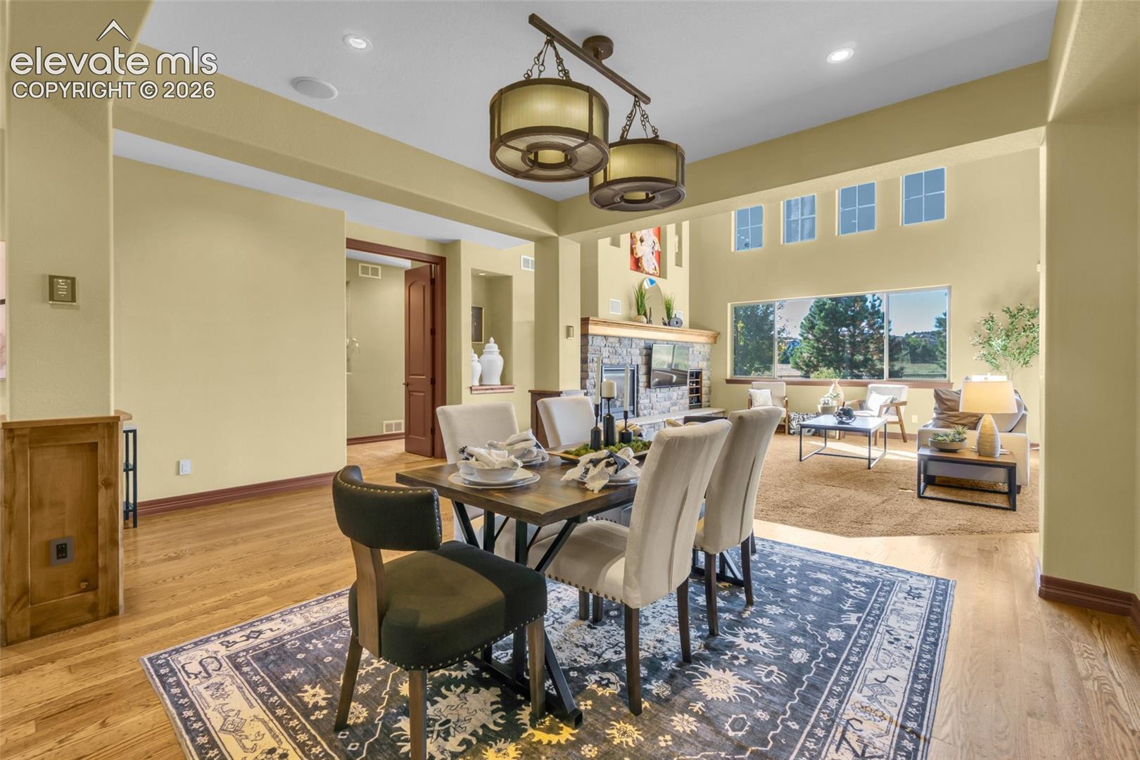 Open-concept space featuring wood-finish flooring, stone-surround fireplace, dual-tier drum pendant lighting, recessed ceiling lights, and transom windows